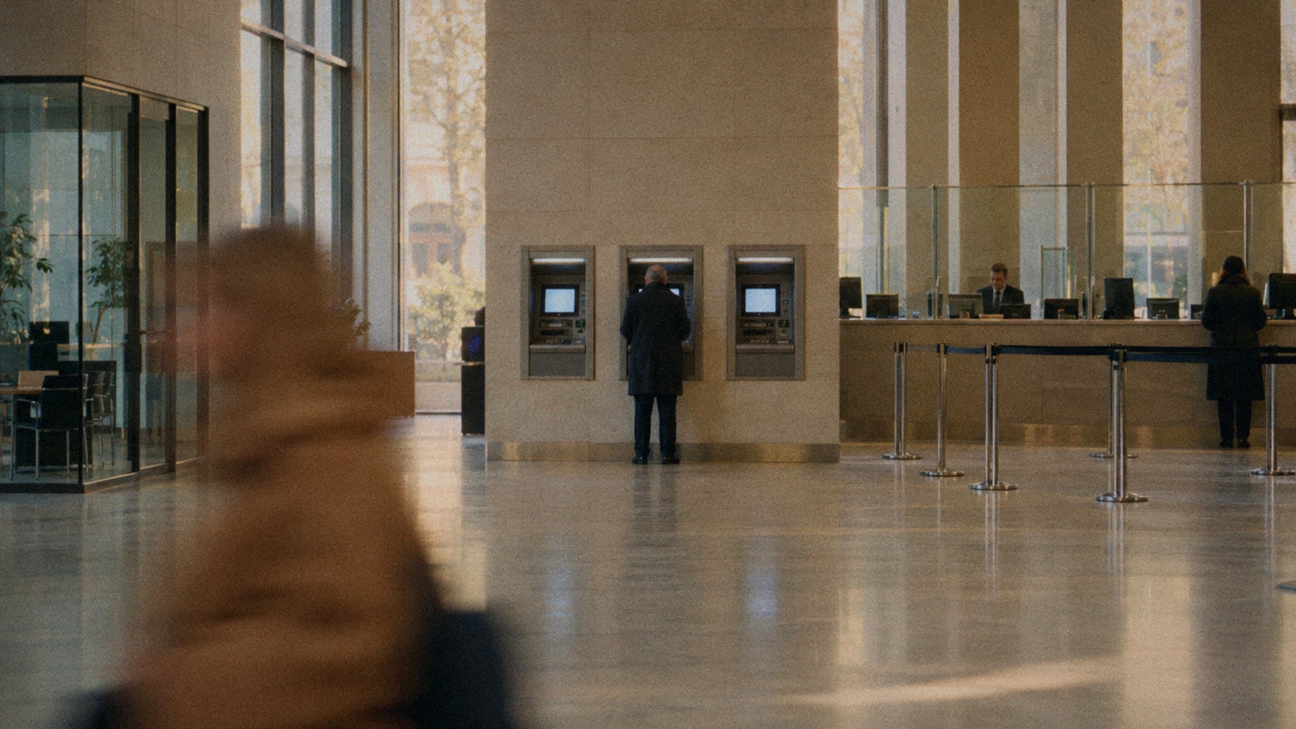 A customer at a cash machine in a bank foyer, with the teller counter beside him.