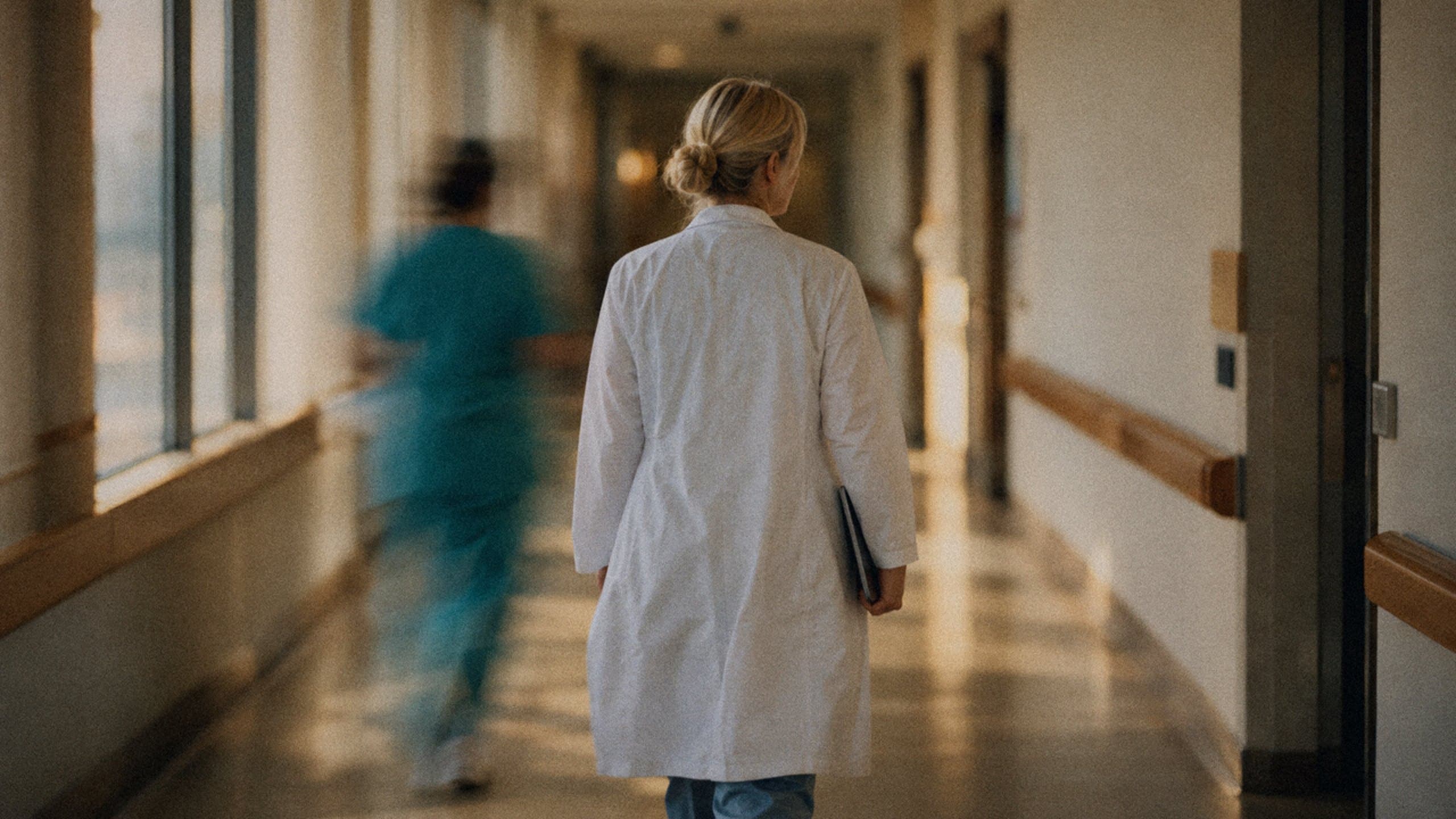 A doctor walking down a calm hospital corridor as a nurse hurries past.