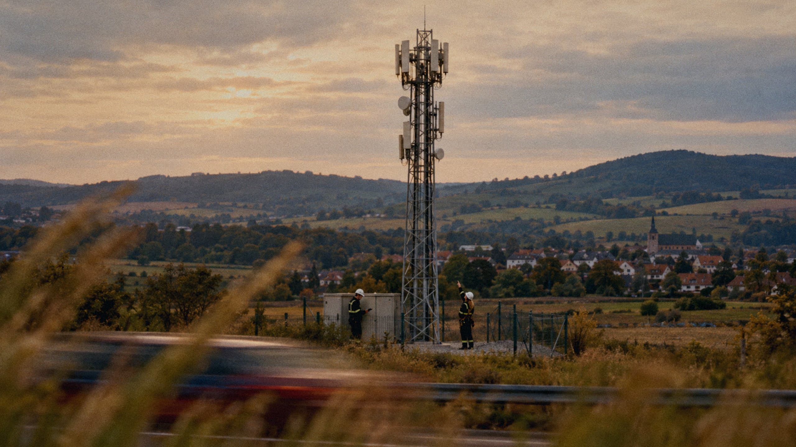 Two telecom engineers at the foot of a mobile base station mast outside a town, a car passing in a blur.