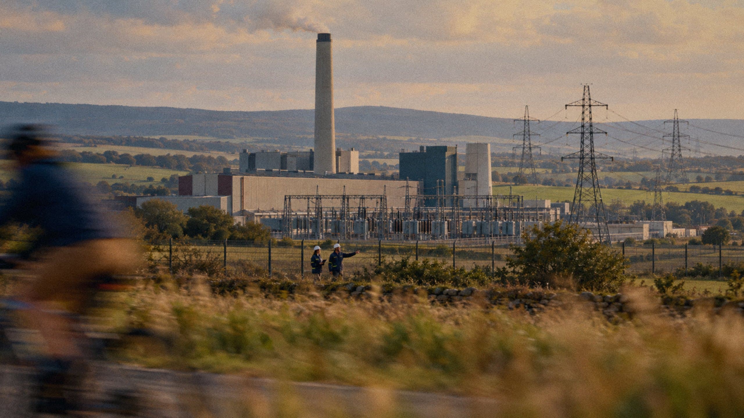 Two engineers at an electricity power station, with high-voltage lines running across the fields.